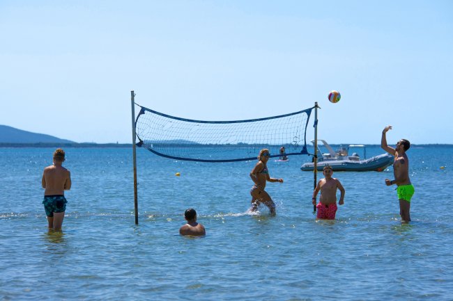 Persone giocano a pallavolo in acqua bassa con rete tesa. Barche sullo sfondo.