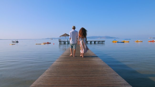 Coppia che cammina su un pontile verso una capanna sul mare, con pedalò in lontananza.