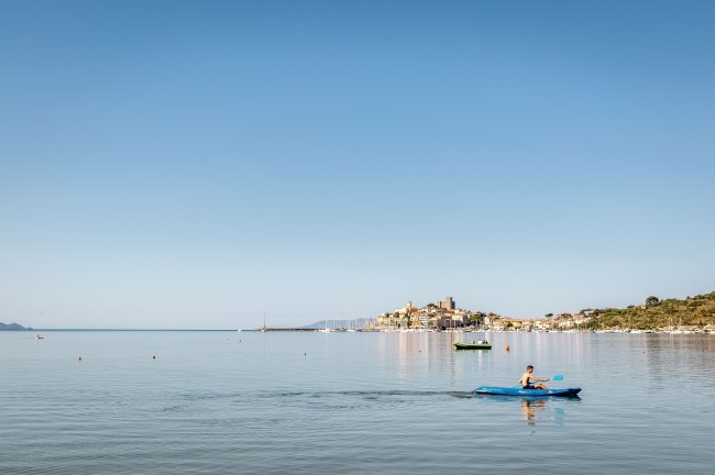 Persona in kayak azzurro su un mare calmo, con un villaggio costiero sullo sfondo.
