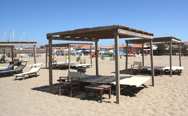 Pagode di legno e lettini su una spiaggia sabbiosa con case colorate sullo sfondo.