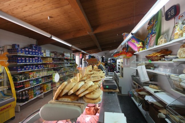 Interno di negozio alimentari con scaffali pieni e pane su bancone.