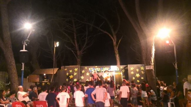 Persone che partecipano a un evento serale all'aperto con luci e palco tra gli alberi.