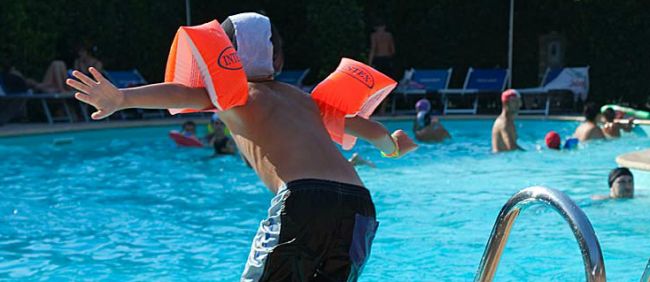 Bambino con braccioli arancioni pronto a tuffarsi in piscina affollata di nuotatori.