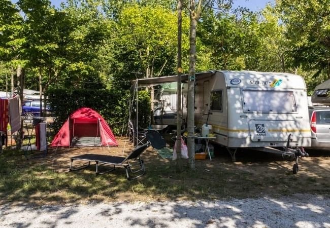 Un'area campeggio con roulotte, tenda rossa, sdraio e vegetazione circostante.