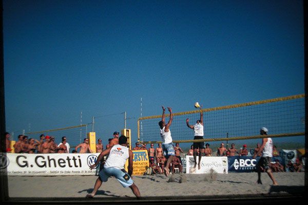 Giocatori di beach volley in azione sulla sabbia, pubblico sullo sfondo.
