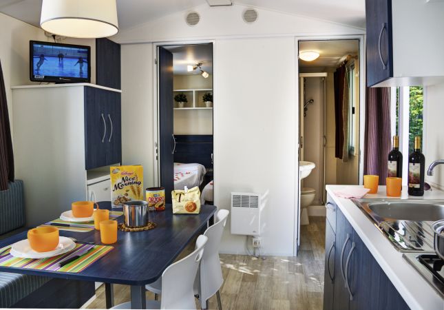 Interno di un camper con tavolo apparecchiato, cucina e TV accesa.