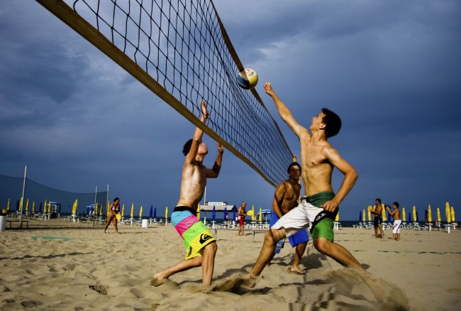 Partita di beach volley su una spiaggia con tre giocatori sotto un cielo nuvoloso.