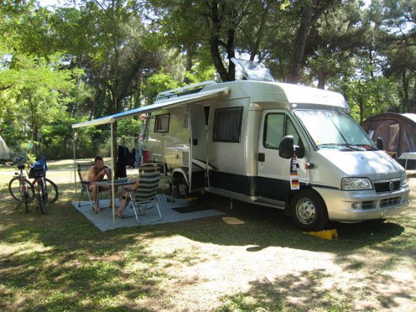 Camper parcheggiato in un'area ombrosa, con due persone sedute a un tavolo sotto un tendalino.