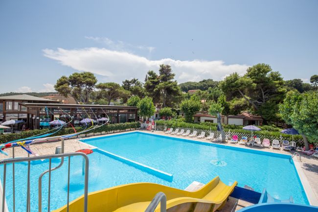 Piscina all'aperto con scivoli e sedie a sdraio, circondata da alberi.