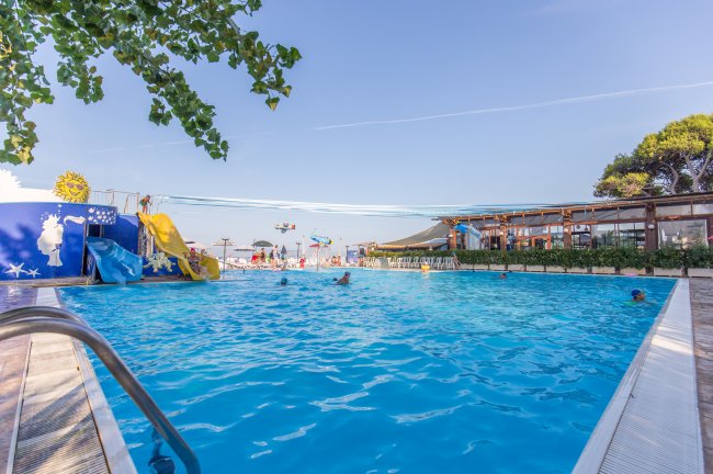 Una piscina con scivoli gialli, persone che nuotano e sedie a sdraio sullo sfondo.