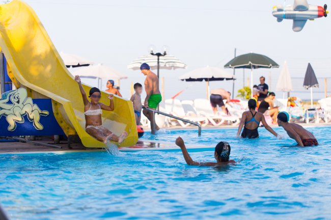 Bambini giocano in piscina con uno scivolo giallo, sotto ombrelloni bianchi e neri.