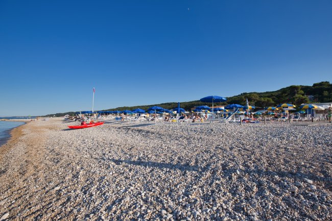 Spiaggia di ciottoli con ombrelloni blu e gialli e un mare calmo sullo sfondo.