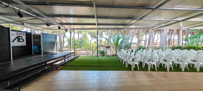 Sala con sedie bianche in plastica e un palco. Sullo sfondo, una pineta e spazi aperti.