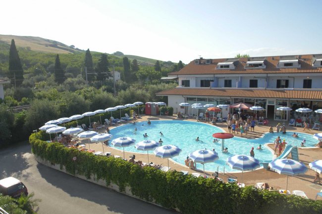Una piscina circondata da lettini e ombrelloni, con un edificio e colline sullo sfondo.