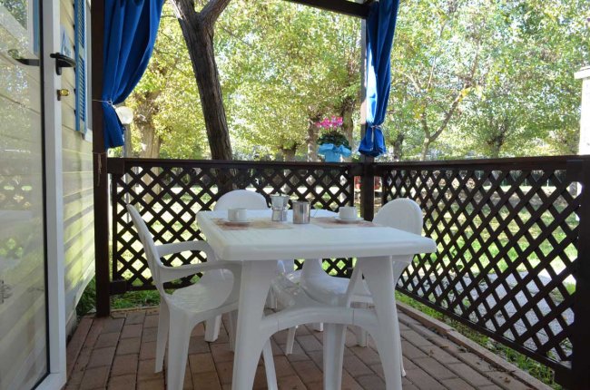 Tavolo bianco con sedie in plastica su una veranda con vista su un giardino alberato.