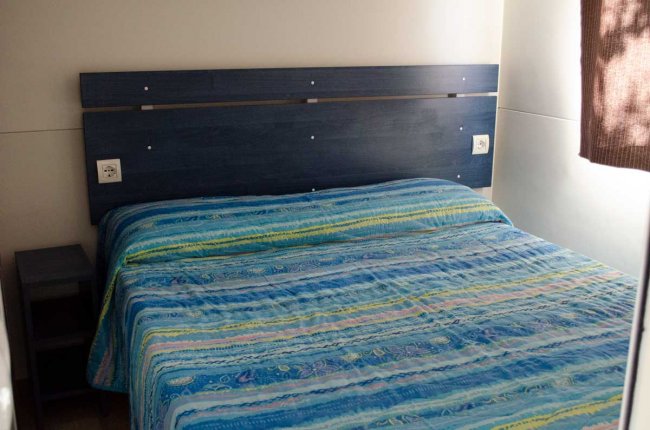 Letto matrimoniale con copriletto blu, testiera in legno, tenda marrone e piccola mensola.