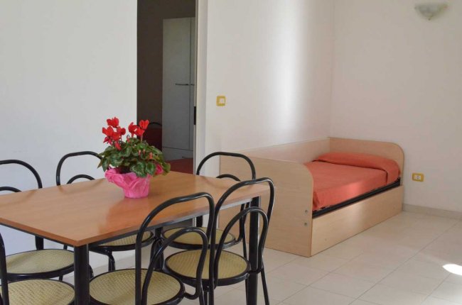 Stanza luminosa con tavolo di legno, sedie nere e un letto singolo con coperta arancione.