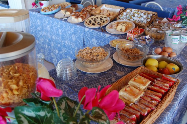 Buffet della colazione con cereali, dolci, frutta e uova esposto su tovaglie blu.