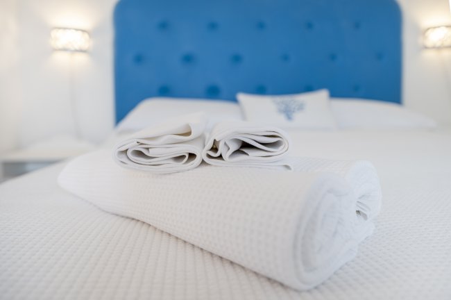 Letto con testiera blu e asciugamani bianchi piegati sopra. Sfondo bianco e luminoso.