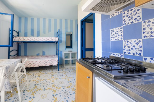 Cucina con fornelli, tavolo con sedie e letti a castello, pareti azzurre e piastrelle colorate.