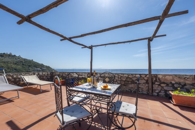 Terrazza con tavolo e sedie, vista mare, sotto pergola di legno.