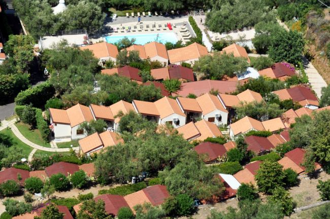 Vista aerea di bungalow con tetti rossi circondati da alberi e una piscina con sdraio.