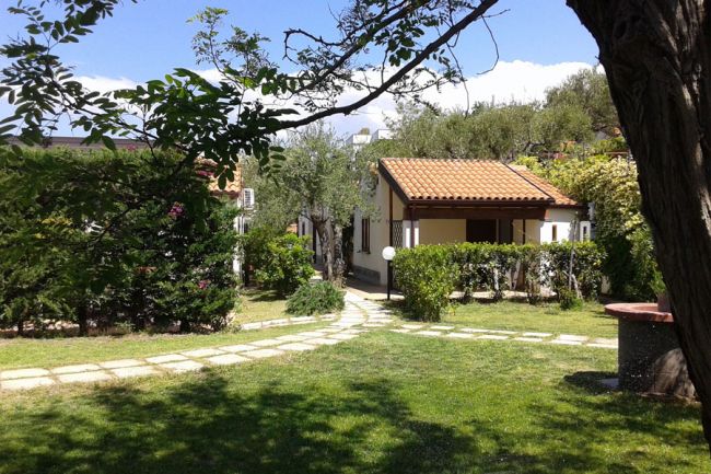 Un giardino con un piccolo edificio dal tetto rosso, sentiero in pietra e alberi ombrosi.