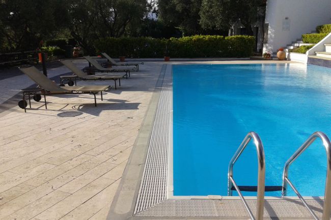 Bordo piscina con sdraio nere, scaletta metallica e vegetazione sullo sfondo.