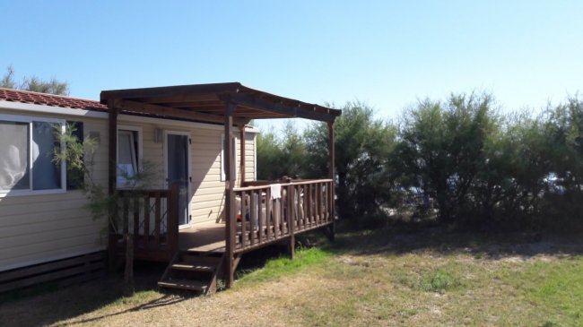 Un bungalow con veranda in legno, circondato da arbusti sotto un cielo sereno.