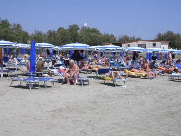 Spiaggia affollata con lettini blu e ombrelloni bianchi, persone che prendono il sole.