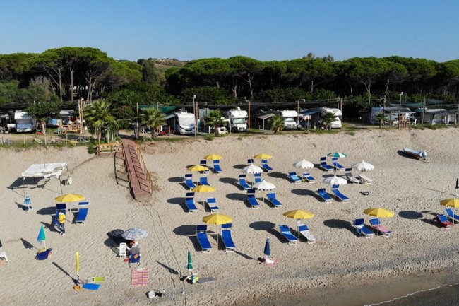 Spiaggia con ombrelloni gialli e lettini blu, alberi e camper sullo sfondo.