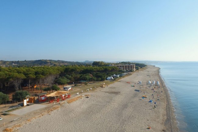 Ampia spiaggia con pochi ombrelloni, lungo un tratto di costa alberato e soleggiato.