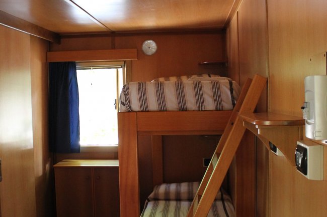 Camera con letti a castello, pareti in legno, piccolo finestra con tenda blu.