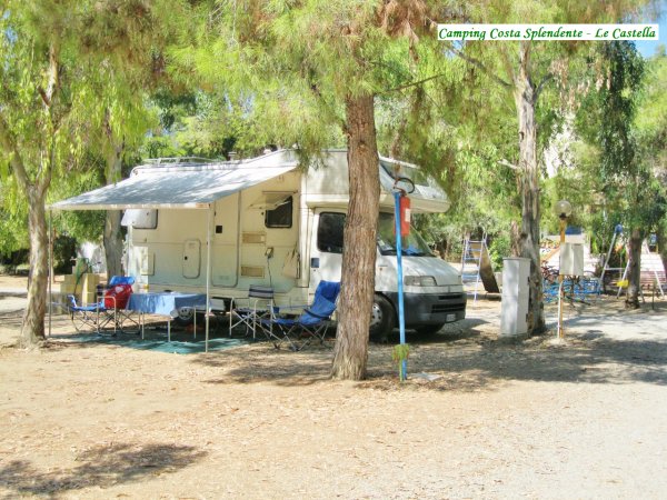 Un camper parcheggiato con sedie e tavolo sotto un telone, circondato da alberi.
