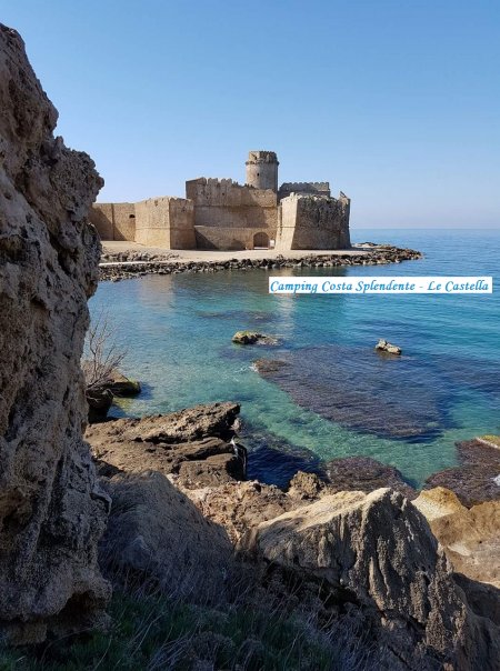 Vista di un antico castello sul mare con acqua cristallina e rocce in primo piano.