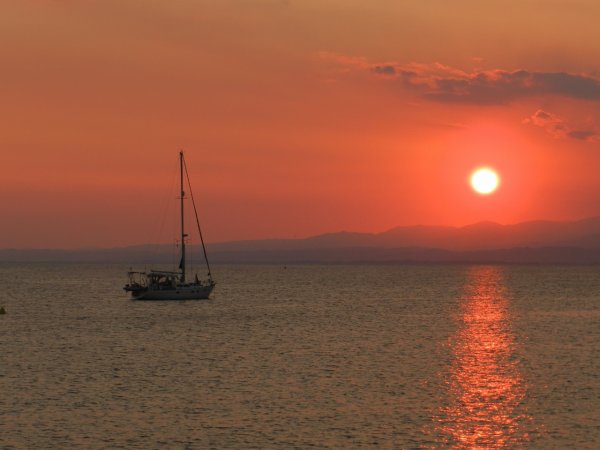 Barca a vela su mare calmo al tramonto, con sole arancione e montagne sullo sfondo.