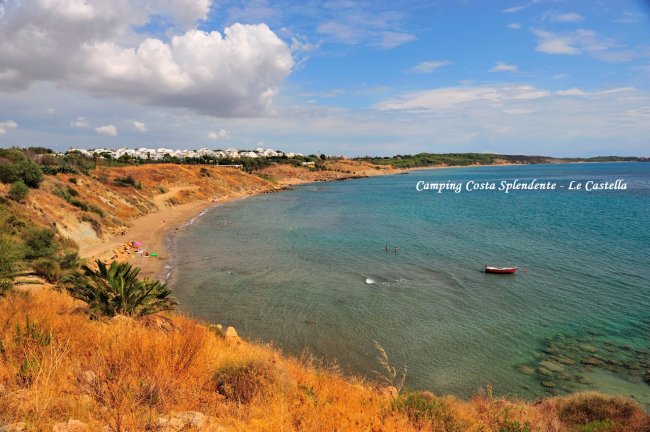 Vista panoramica di una spiaggia con scogliere, vegetazione e case bianche in lontananza.