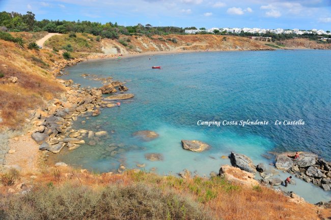 Baia rocciosa con mare turchese, vegetazione secca e un piccolo sentiero.