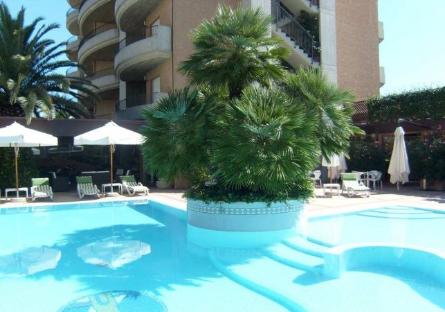 Piscina con palme centrali, ombrelloni bianchi e edificio sullo sfondo.