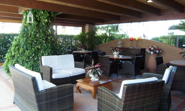 Un salotto esterno con mobili in vimini sotto una tettoia, decorato con piante.