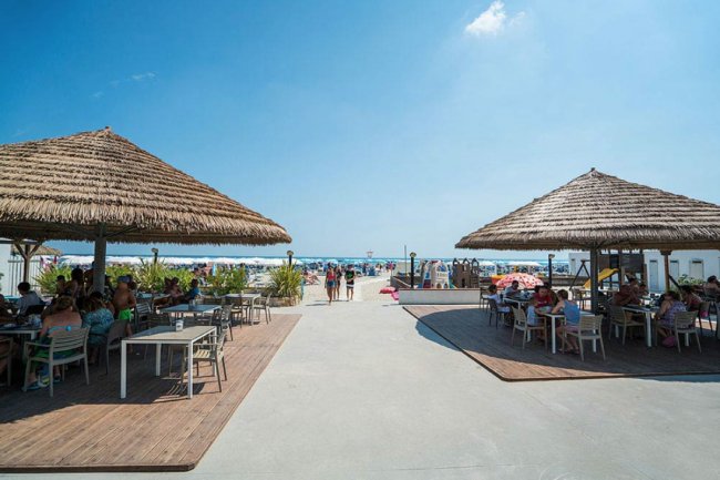 Due tettoie di paglia su una terrazza fronte mare con tavoli e persone sedute.