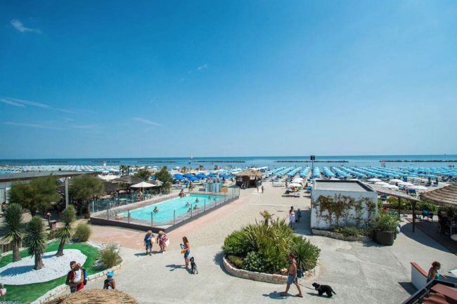 Area balneare con piscina, spazi verdi e lettini sotto ombrelloni azzurri.