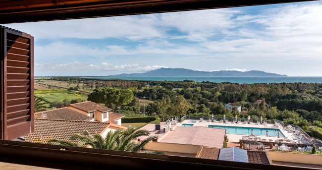 Vista panoramica su una piscina, tetti di case, paesaggio verde e mare in lontananza.