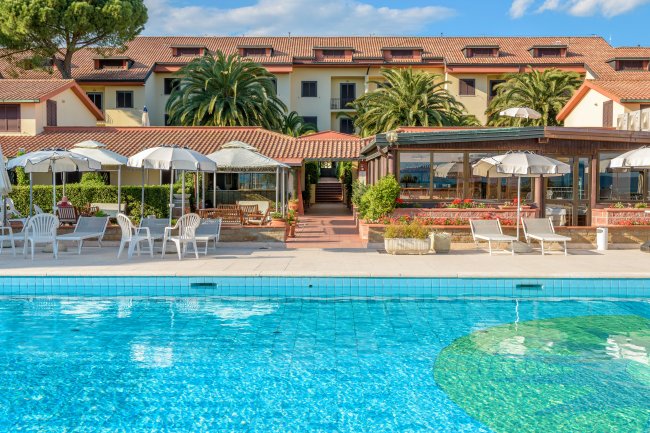 Piscina con lettini e ombrelloni davanti a un edificio con tetti rossi e palme.