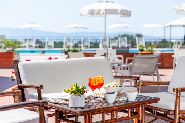 Tavolo esterno con aperitivi, sedie in legno e vista su piscina e ombrelloni bianchi.