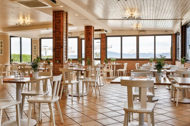 Ristorante luminoso con sedie e tavoli bianchi. Ampie finestre e piante decorative.