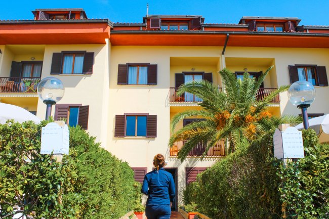 Persona cammina verso un edificio a tre piani con balconi e una palma in giardino.