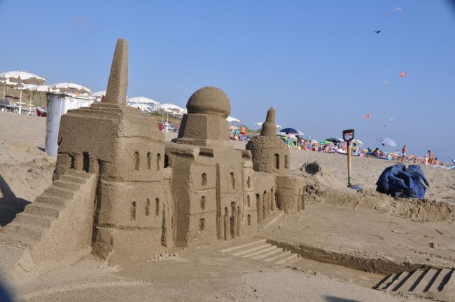 Castillo di sabbia dettagliato su una spiaggia affollata, con ombrelloni sullo sfondo.