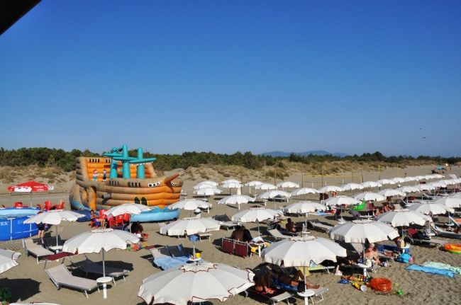 Spiaggia estiva con lettini e ombrelloni bianchi, più un grande gonfiabile a forma di nave.