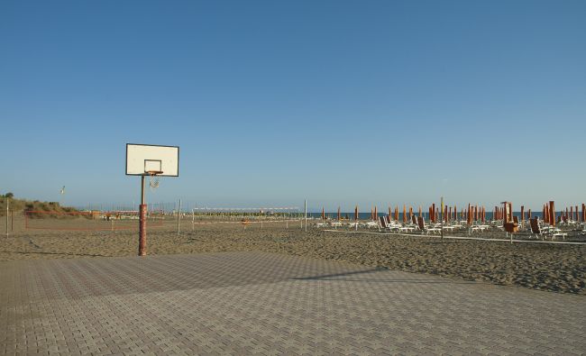 Campo da basket su una spiaggia con ombrelloni chiusi e mare all'orizzonte.